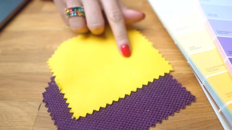 A Close-Up of A Person with Red Nail Polish Pointing to Vibrant Yellow and Purple Fabric Swatches, with A Color Palette Guide on The Side, Illustrating the Concept of Color Psychology in Living Room Decor